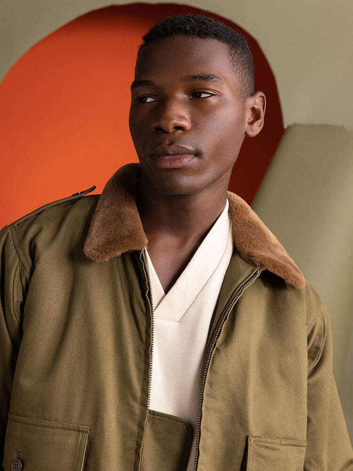 A person wearing the 1940s B-10 Bomber Jacket in olive by Alpha Industries, featuring a brown collar, gazes to the side.