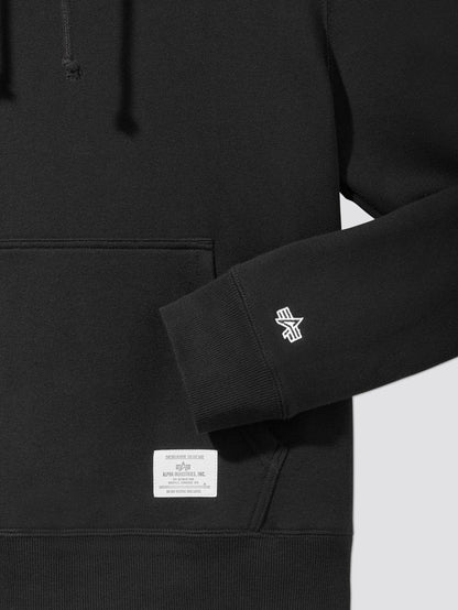 Close-up of the Black Essential Hoodie, a True Wardrobe Staple, Showcasing a Front Pocket, Logo on the Sleeve, and an Alpha Industries Label with Text Above the Pocket.