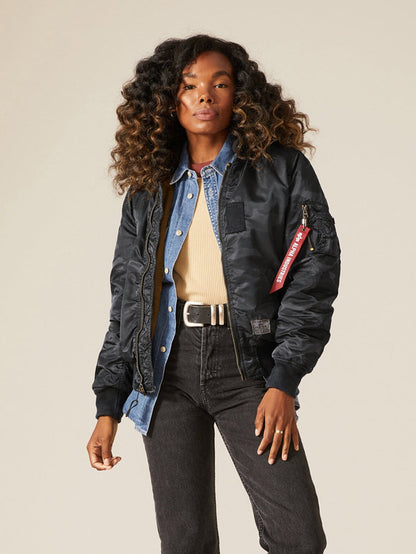 A person wearing an Alpha Industries MA-1 Hooded Battlewash Bomber Jacket in Black Woodland Camo over a denim jacket and beige shirt, paired with dark jeans.