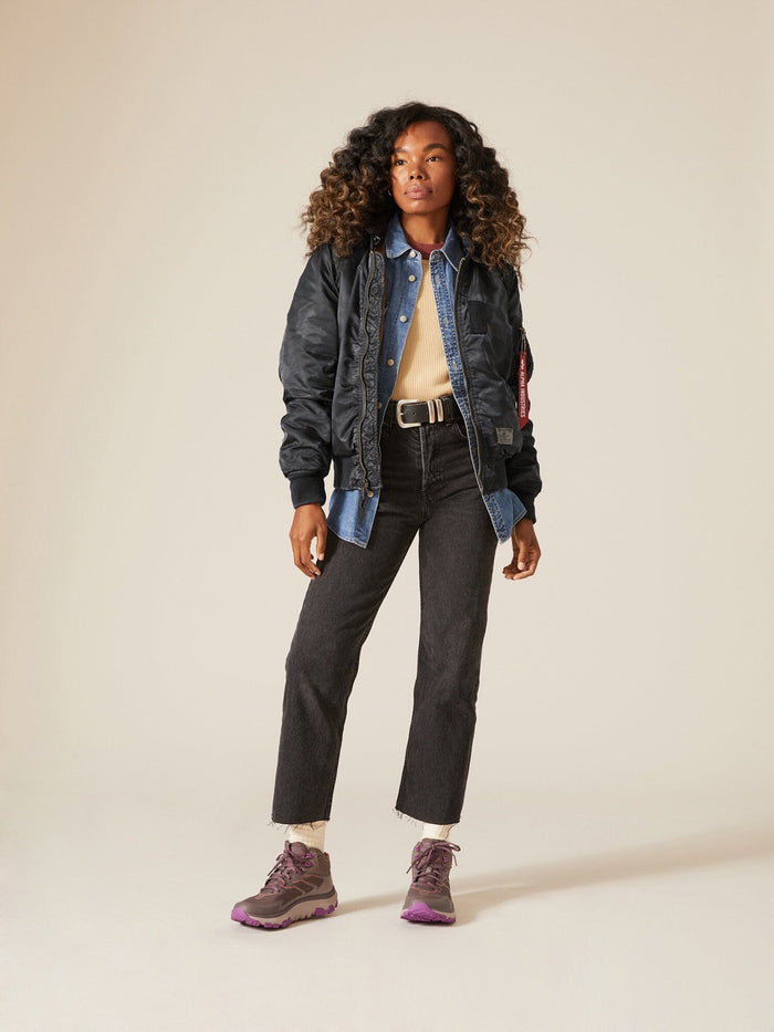 A person stands confidently wearing an Alpha Industries MA-1 Hooded Battlewash Bomber Jacket, denim shirt, light sweater, dark pants, and purple sneakers.