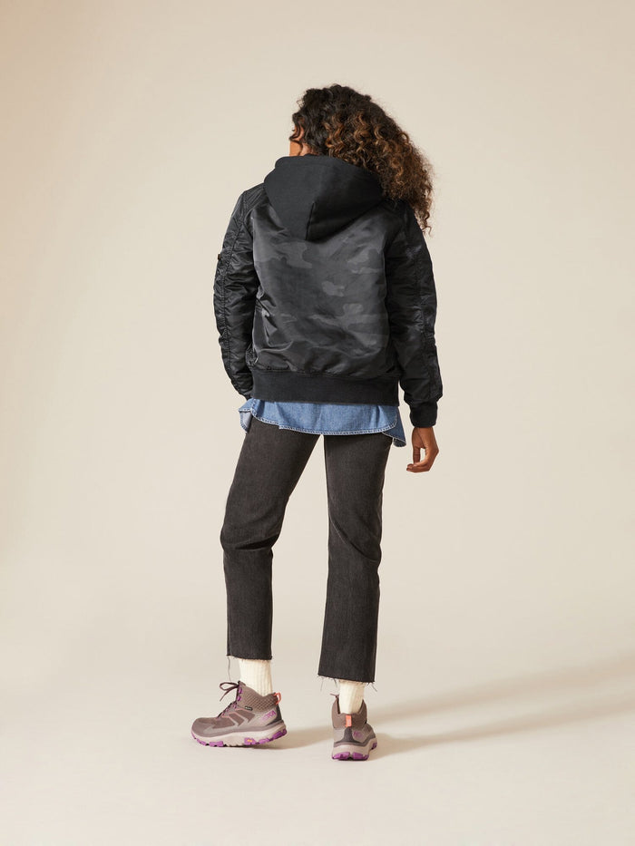 A person with curly hair faces away, wearing an Alpha Industries MA-1 Hooded Battlewash Bomber Jacket, a denim shirt, black pants, and pink sneakers.